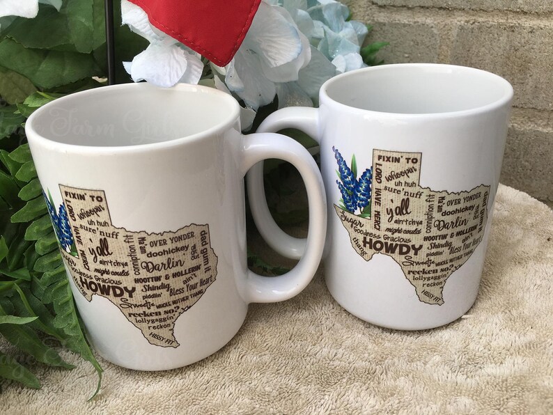 May include: Two white ceramic mugs with a Texas-shaped design. The design includes the words "Howdy", "Y'all", and "Darlin'", along with a bluebonnet flower illustration. The mugs have a curved handle.
