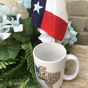 May include: A white ceramic mug with a Texas-shaped graphic featuring the words "Howdy" and "Y'all." A small Texas flag with a white star and red and blue stripes is displayed next to the mug. The mug is surrounded by greenery and blue flowers.