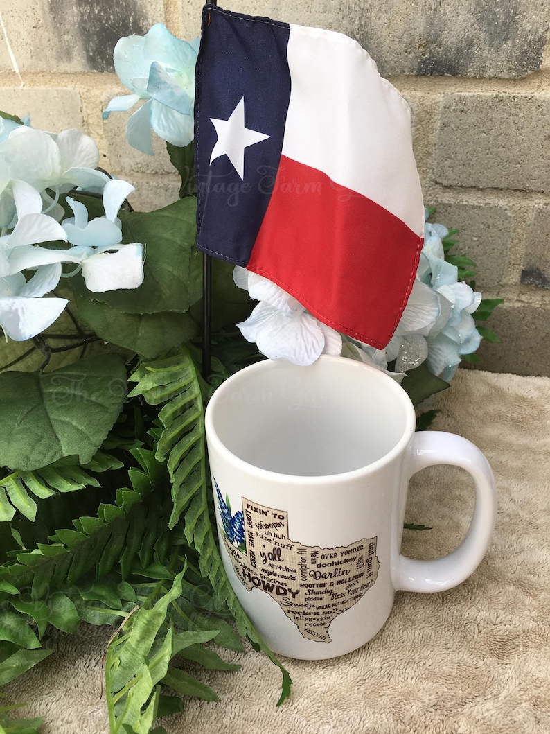 May include: A white ceramic mug with a Texas-shaped graphic featuring the words "Howdy" and "Y'all." A small Texas flag with a white star and red and blue stripes is displayed next to the mug. The mug is surrounded by greenery and blue flowers.