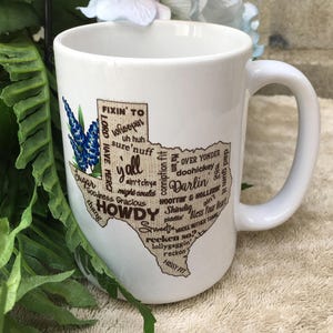 May include: White ceramic mug featuring a Texas-shaped design with the word "Howdy" and other Texas slang terms. The design includes a bluebonnet flower illustration. The mug has a large handle.