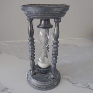 May include: A gray hourglass with a clear glass center filled with white sand. The hourglass is supported by a decorative, ornate gray metal frame. The top and bottom are circular, and the frame has a detailed design.