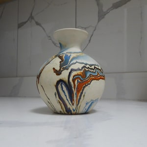 May include: A ceramic vase with a rounded body and a slender neck. The vase is mainly white with a marbled pattern in blue, orange, and brown. The rim of the vase is a solid off-white colour.