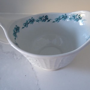 May include: White ceramic gravy boat with a blue floral design around the rim. The boat has a textured, woven pattern on the bottom.