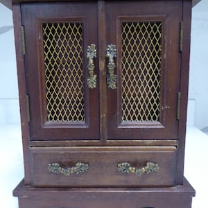 May include: A dark brown wooden jewelry box with two doors and a drawer. The doors have a gold wire mesh insert and ornate brass handles. The drawer has two ornate brass handles.