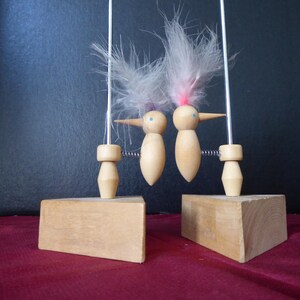 May include: Two wooden bird figurines with feather crests and blue eyes are connected by springs and suspended from thin rods. The birds sit on wooden blocks, with a dark blue background and a red surface.