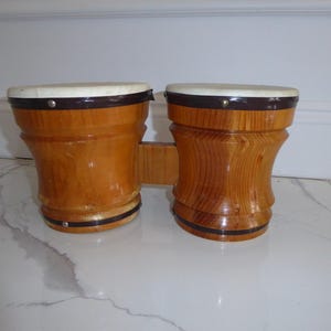 May include: A pair of wooden bongos with light brown wood and white drumheads. The drums are connected by a wooden bridge. The rims are dark brown. The bongos are sitting on a white and gray tiled surface.