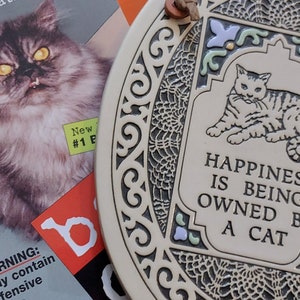 May include: A white ceramic plaque with a decorative border and a cat illustration. The plaque reads "Happiness is being owned by a cat."
