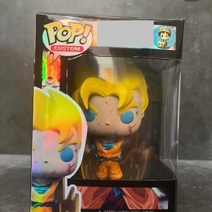 Pode incluir: Uma figura Pop! personalizada de um personagem de anime com cabelo amarelo, olhos azuis e uma cicatriz no rosto. A figura está vestindo um macacão laranja e está em pé sobre uma base vermelha e laranja. A figura está em uma caixa de plástico transparente com um logotipo Pop! preto e branco na frente. A caixa também tem um rótulo de advertência sobre os perigos de asfixia.