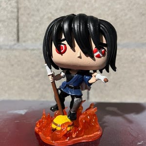May include: A custom-painted Funko Pop figure of a character with black hair, red eyes, and a blue and black outfit. The figure is holding a brown staff and standing on a yellow and orange base with flames.