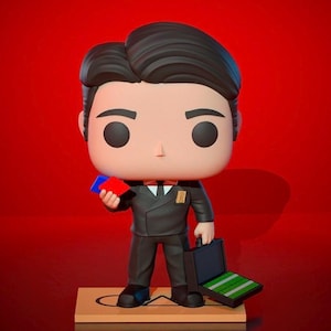 Peut inclure: Une figurine en vinyle noir et blanc d'un homme en costume noir tenant une carte rouge et bleue et une mallette noire avec un motif vert et blanc à l'intérieur. La figurine est debout sur une plateforme brune avec un cercle blanc.