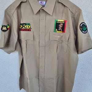 Puede incluir: Camisa caqui de manga corta con múltiples parches y el texto "Bob Marley". La camisa tiene dos bolsillos delanteros y charreteras. La talla es XL.
