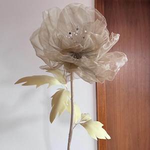 May include: A large, artificial flower with a beige, translucent petal design. The flower has a long, light brown stem with pale yellow leaves. Decorative accents are visible in the center of the flower. The flower is set against a white wall and a wooden door.