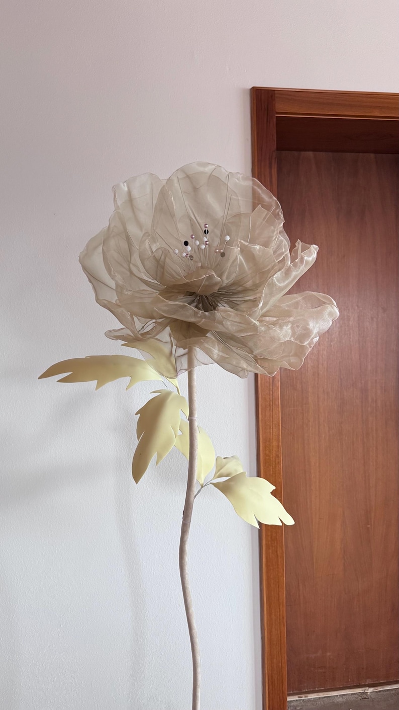 May include: A large, artificial flower with a beige, translucent petal design. The flower has a long, light brown stem with pale yellow leaves. Decorative accents are visible in the center of the flower. The flower is set against a white wall and a wooden door.