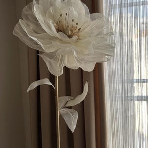 May include: A large, off-white artificial flower with delicate, layered petals and a golden center. The flower is mounted on a slender, gold-colored stem with fabric leaves. The backdrop includes neutral-toned curtains and a window.