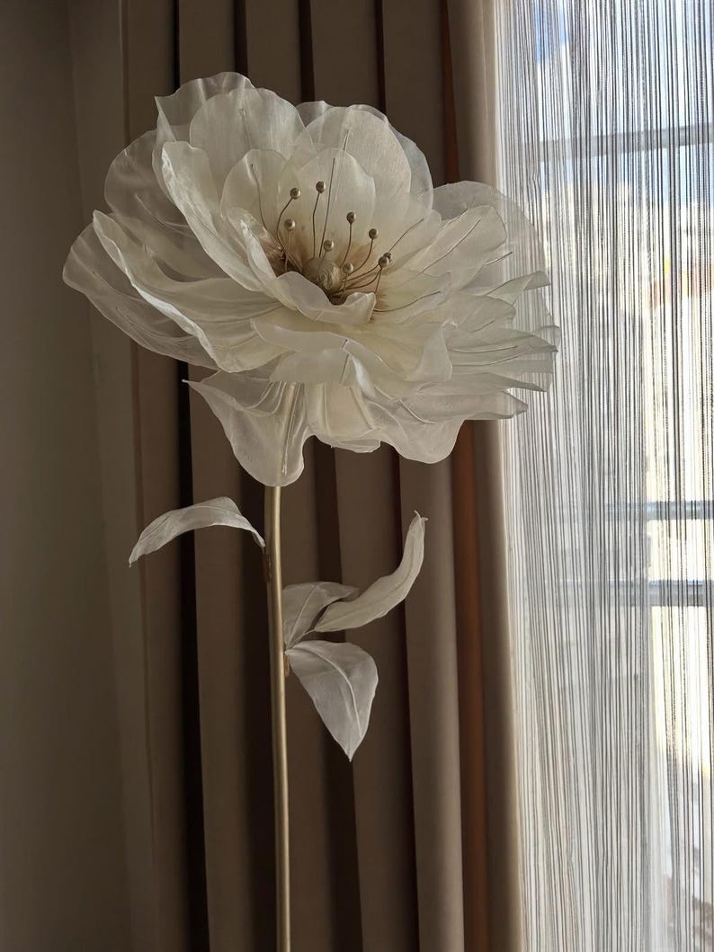 May include: A large, off-white artificial flower with delicate, layered petals and a golden center. The flower is mounted on a slender, gold-colored stem with fabric leaves. The backdrop includes neutral-toned curtains and a window.