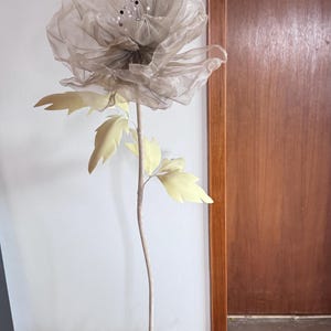 May include: A large, artificial flower with a sheer, gray petal design and a stem. The flower has light yellow leaves and is set in a green, moss-covered base. The stem is a light brown color.