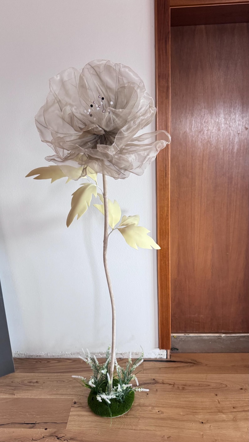 May include: A large, artificial flower with a sheer, gray petal design and a stem. The flower has light yellow leaves and is set in a green, moss-covered base. The stem is a light brown color.