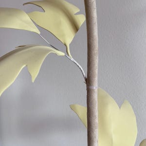May include: Close-up of a decorative artificial plant with pale yellow leaves and a textured, light brown stem. The leaves are attached to the stem with thin, silver-colored wire.