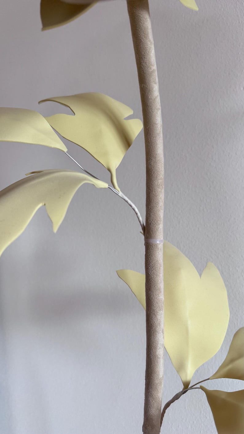 May include: Close-up of a decorative artificial plant with pale yellow leaves and a textured, light brown stem. The leaves are attached to the stem with thin, silver-colored wire.
