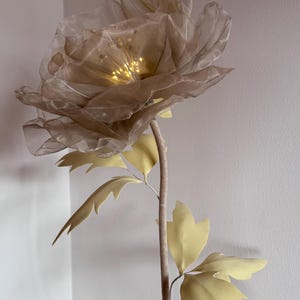 May include: A large, artificial flower with a beige, translucent petal design and a warm, glowing center. The stem is a light brown color, with pale yellow leaves. The flower is a decorative accent piece.