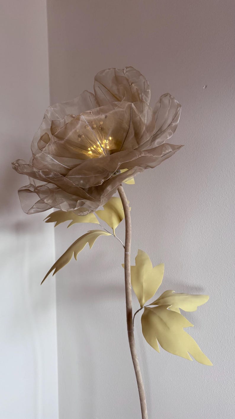 May include: A large, artificial flower with a beige, translucent petal design and a warm, glowing center. The stem is a light brown color, with pale yellow leaves. The flower is a decorative accent piece.