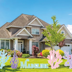 May include: Lawn decorations spelling out "Madeline" with coral and seaweed designs in shades of blue, purple, and pink. The decorations are placed on a green lawn in front of a house.