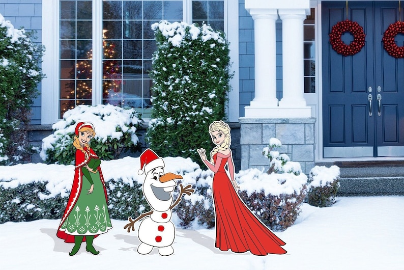May include: A snowman wearing a Santa hat stands next to two girls in winter outfits. The girl on the left is wearing a red and green dress with a red cape. The girl on the right is wearing a red dress. The scene is set in front of a house with a blue door and a snowy yard.