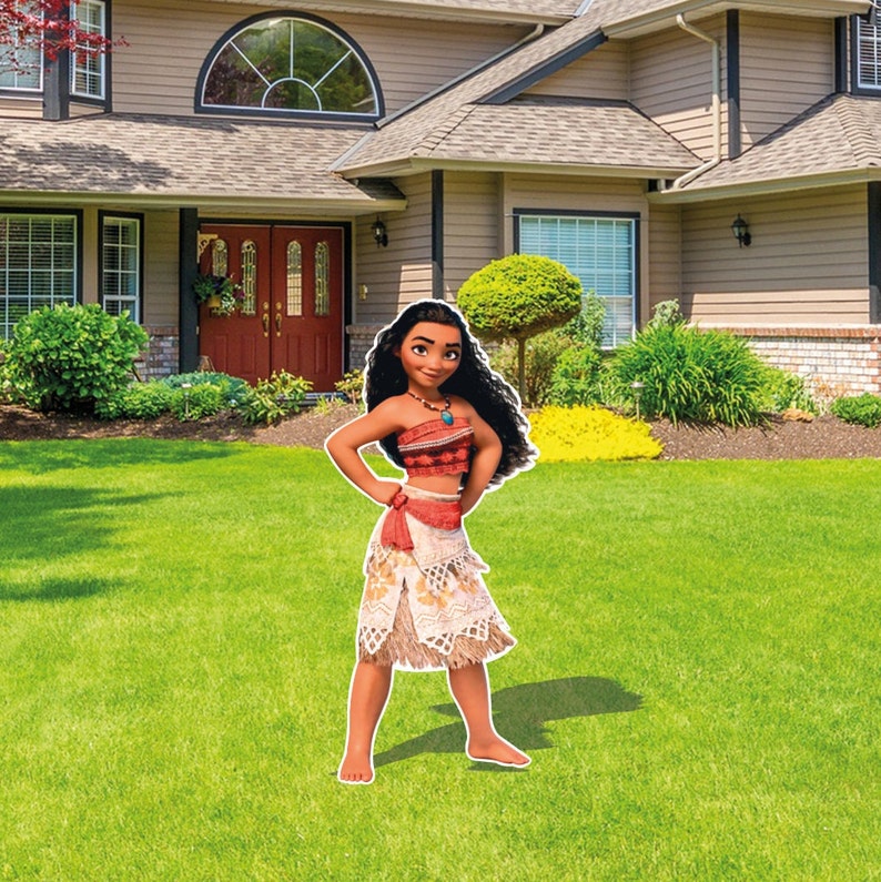 May include: A life-sized cutout of a character with long dark wavy hair, wearing a red patterned crop top, a beige skirt with a red sash, and a blue stone necklace. The figure stands with hands on hips, positioned on a green lawn in front of a house.