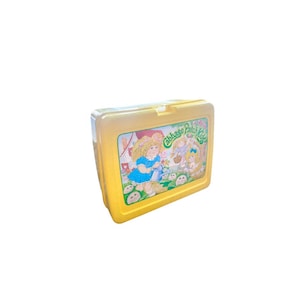 Op de afbeelding: Gele plastic lunchbox met een Cabbage Patch Kids-ontwerp. De lunchbox heeft een afbeelding van drie Cabbage Patch Kids-poppen met bloemen en een groene achtergrond.
