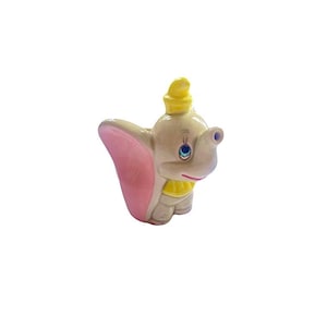 May include: A ceramic Dumbo the elephant figurine. The figure has a light beige body, large pink ears, and a yellow hat and collar. The eyes are blue, and the trunk is extended. This collectible figurine is a popular character.
