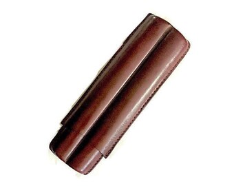 May include: A brown leather cigar case with a two-cigar capacity. The case is rectangular with rounded edges and a stitched border. The leather has a smooth, polished finish. The case is designed to protect and carry cigars.