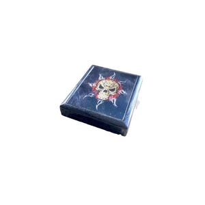 Puede incluir: Una caja rectangular negra con un diseño de calavera y rayos de sol en la tapa. La calavera es dorada con detalles rojos, y los rayos de sol son blancos. La caja tiene un cierre plateado.