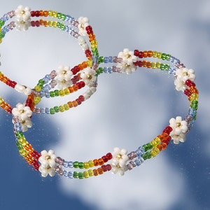 May include: A rainbow beaded bracelet with white flower accents. The bracelet is made of small, colorful beads and features a repeating pattern of white flower charms.
