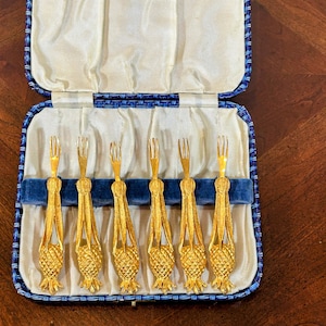 May include: A set of six gold-colored dessert forks in a blue and cream-colored presentation box. The forks have a pineapple design on the handles and are arranged neatly in a velvet-lined case. The case is open, revealing the forks.