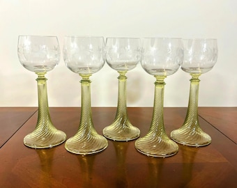 Puede incluir: Cinco copas de vino transparentes con diseños florales grabados en los cuencos. Los tallos son de color verde claro con un diseño retorcido. Las copas se exhiben sobre una superficie de madera marrón oscuro.