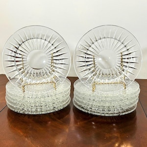 May include: Two stacks of clear glass plates with a starburst design, each plate supported by a gold-colored metal stand. The plates are arranged on a dark wooden surface, showcasing a classic tableware set.