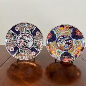 May include: Two decorative ceramic plates with intricate Japanese-style designs. One plate features a floral motif, while the other showcases a landscape scene with a pagoda and mythical creatures. Both plates have a dark blue border.
