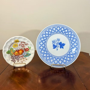 May include: Two decorative ceramic plates on display stands. The plate on the left has a colorful fruit and floral design. The plate on the right has a blue and white floral pattern.