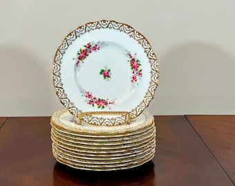 Puede incluir: Una pila de 10 platos de cena de porcelana blanca con un borde dorado y un diseño floral. Los platos presentan un delicado patrón floral con rosas rosas y hojas verdes.