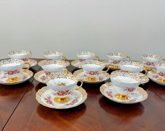 Puede incluir: Un juego de diez tazas de té y platillos blancos con un patrón floral y ribete dorado. Las tazas y los platillos están dispuestos en dos filas sobre una superficie de madera.