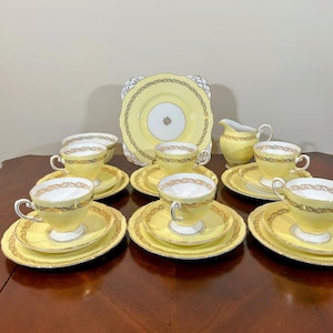 Puede incluir: Un juego de té amarillo vintage con detalles dorados, que incluye tazas, platillos, platos, una jarra de crema y una fuente cuadrada. El juego está dispuesto sobre una superficie de madera oscura, mostrando un diseño clásico.