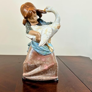 May include: A ceramic figurine of a girl holding a white goose. The girl has brown hair and is wearing a blue and green dress with a pink skirt. The goose has a long neck and is facing the girl. The figurine is on a wooden surface.