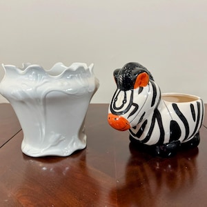 May include: Two ceramic planters, one white with a floral design and the other shaped like a zebra with black and white stripes and orange accents.