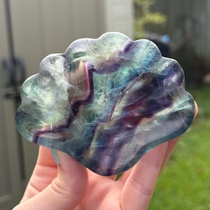 May include: A multi-colored fluorite crystal carving in a scalloped shape. The crystal features shades of purple, blue, green, and pink. The translucent stone is held in a hand against a blurred green background.