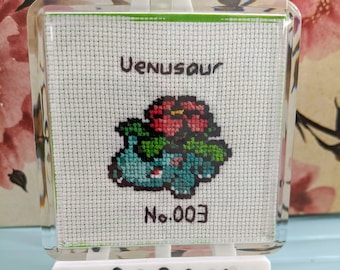 Op de afbeelding: Een kruissteekkunstwerk van Venusaur, een groen en blauw Pokémon met een rode bloem op zijn hoofd. Het kunstwerk is ingelijst in een doorzichtig plastic frame en wordt getoond op een wit ezel. Het kunstwerk is gelabeld met "Venusaur" en "No. 003".