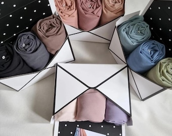 Op de afbeelding: Verschillende opgerolde hijabs in diverse kleuren, waaronder zwart, grijs en tinten roze en blauw, worden getoond in witte geometrische dozen met zwarte stippen. Elke hijab is netjes opgerold en vastgezet, met een verscheidenheid aan zachte, effen kleuren.