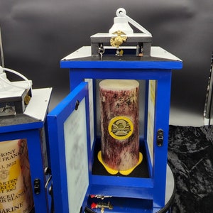 May include: Two blue wooden candle lanterns with glass windows. The lantern on the right has a white candle with a gold emblem inside. The lantern on the left has a white background with black text and a gold emblem. The text reads "HONY FRANK 3, 2022 ORTBN KE CO T 3062 MCRD RIS ISLAND MARINE 201".
