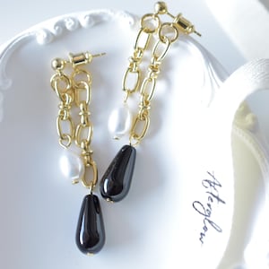 Peut inclure: Boucles d'oreilles à chaîne dorée avec des perles blanches et des accents de goutte d'eau noirs. Les boucles d'oreilles ont une fermeture à tige. Les boucles d'oreilles sont présentées sur une surface blanche.