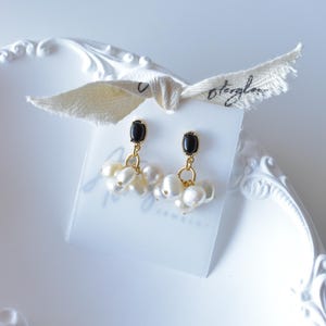 May include: A pair of earrings featuring a black stone and a cluster of white pearls, suspended from gold-coloured metal. The earrings are presented on a white card, tied with a cream ribbon. The ribbon has handwritten text.