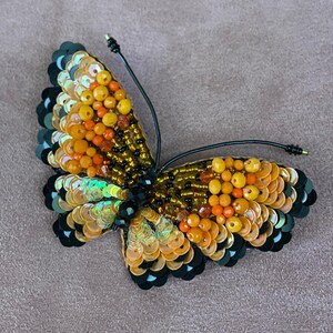 May include: A beaded butterfly brooch with black, orange, and yellow sequins. The butterfly has a black wire attached to the back for pinning.